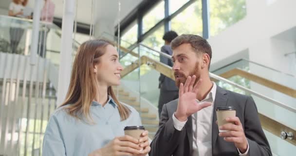 In der Pause stehen männliche und weibliche Mitarbeiter mit Kaffeetassen aus Pappmaché im Büro und unterhalten sich. schöner männlicher Arbeiter spricht zu seiner Kollegin und sie sieht erstaunt aus. — Stockvideo