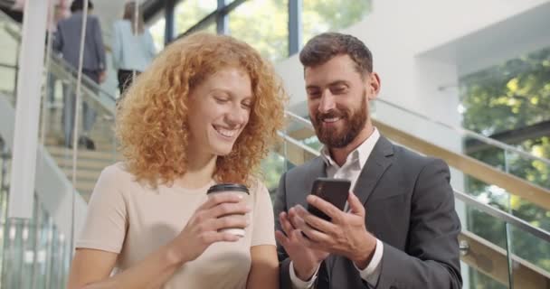 Hansome office worker in 30s showing funny photos to his female coworker at smartphone. Coworkers standing at hall with cofee cups, using mobilephone and looking at screen. People and tech. — Stock Video