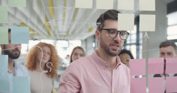 Bonito líder de equipe masculino explicando nova estratégia de projeto para seus colegas em reunião. Empresário na década de 30 usando placa de vidro e conversando com seus subordinados no escritório moderno. Conceito de criatividade . — Vídeo de Stock