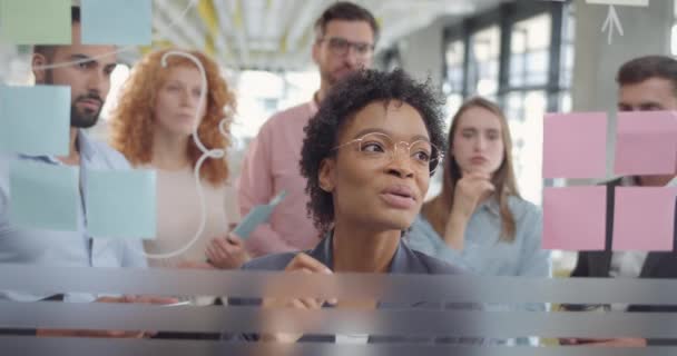 Attraktive afrikanische Geschäftsfrau, die ihren Amtskollegen neue Ideen erklärt. Team junger Start-ups beim Brainstorming mit Glasplatten und klebrigen Papieren. — Stockvideo