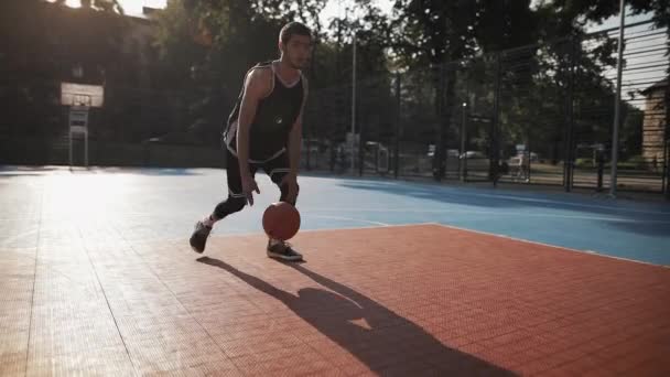 バスケットボール、ハンドリングボール、足の間で跳ね返り、バスケットボールストリートコートでバスケットに投げ込むために白人のスポーティ男の列車の近くのビュー。健康的なライフスタイルとスポーツコンセプト. — ストック動画