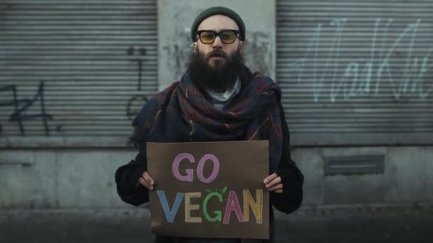 Lviv, Oekraïne - 27 november 2019: De tijd verstrijkt dat een man bord vasthoudt Go Vegan en buiten staat. Hipster man ondersteunt vegetarische eetcampagne. menigte van mensen passeren op de achtergrond. — Stockvideo