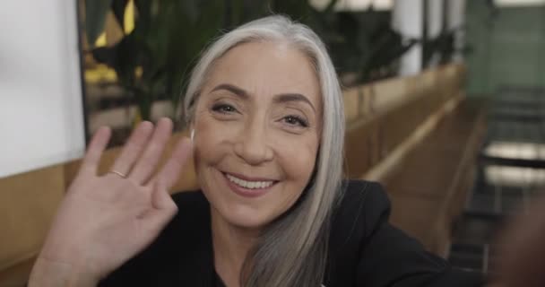 Camera view of smiling mature beautiful lady having video call with friends. Mature businesswoman communicating and vawing to her smartphone camera while sitting in modern cafe. — 비디오