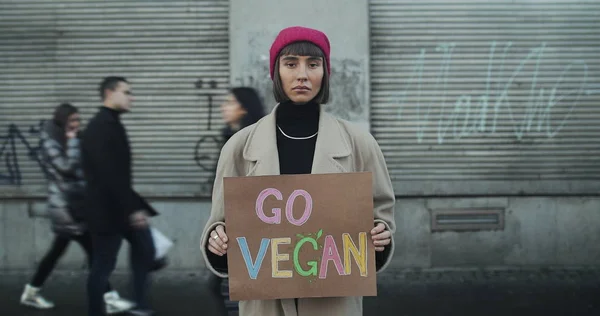 Lviv, Ukraina - 27 November 2019: Gadis hipster muda memegang spanduk bertanda Go Vegan dan berdiri di luar. Waktu berlalu. Orang-orang melewati latar belakang . Stok Lukisan  
