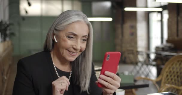 Kulaklıklı zarif bir iş kadını elinde akıllı bir telefon tutuyor ve video görüşmesi yapıyor. Yetişkin modern kadın kafede oturuyor ve torunla video konferans kullanarak iletişim kuruyor.. — Stok video
