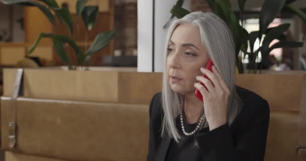 Close up of middle aged beautiful businesswoman talking on smartphone. Concentrated elderly female boss sitting on couch and having phone conversation. Indoor. Concept of business. — Stock Video