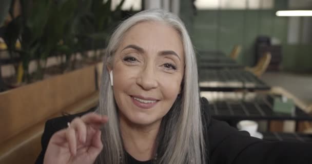 Primer plano de elegante mujer de negocios de mediana edad en auriculares que tienen videocall en el teléfono inteligente. Vista de la cámara de la hermosa anciana que se comunica, utilizando la aplicación de vídeo de su teléfono mientras está sentado en la cafetería . — Vídeos de Stock