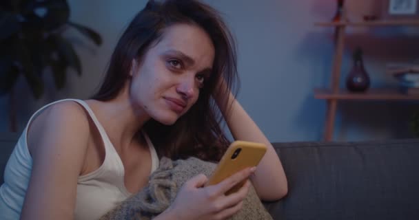 Close-up van depressief meisje zittend op de bank met smartphone in foetale positie. Jonge ongelukkige vrouw met tranen op haar gezicht kijken naar mobiele telefoon scherm en denken aan slecht nieuws. — Stockvideo