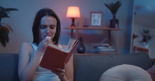 Framifrån på ung upprörd flicka med tårar på hennes ansikte läsa bok. Millennial brunett kvinna sitter på soffan gråter på grund av boklotter och med hjälp av papper. hemmabas. — Stockvideo