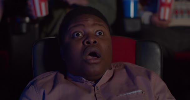 Close up of millennial man sitting in chair and reacting in awe to exciting moment in movie. Guy watching movie in cinema with opened mouth and big eyes and eating popcorn. — Stock Video