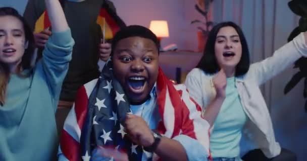 Close up af spændte multietniske venner med flag ser sport på tv og sidder på sofaen. Fokus på afro amerikansk fyr med nationalt flag på skuldre smilende og syngende . – Stock-video