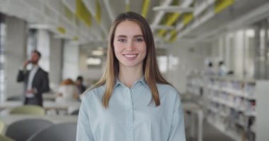 Smiling caucasian girl intern standing at modern office coworking space. Portrait of young female office manager looking to camera and having good mood. Blurred office background.