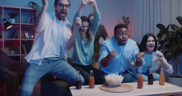 Les gars heureux et les filles célèbrent la victoire de l'équipe tout en étant assis sur le canapé à la maison. Groupe d'amis excités se réjouissant de la victoire tout en regardant des jeux de sport à la télévision. Concept de sport et d'émotions . — Video