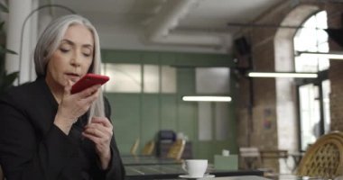 Front view of middle aged woman business owner using smartphone voice commander while sitting in cafe. Modern old lady holding phone and dictating voice message. Concept of people and tech.