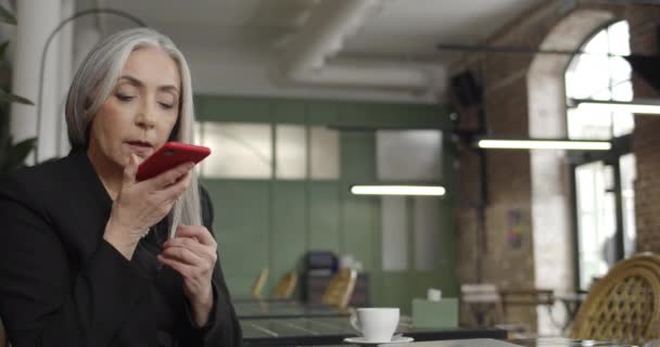 Vooraanzicht van middelbare leeftijd vrouw ondernemer met behulp van smartphone voice commander tijdens het zitten in cafe. Moderne oude dame die telefoon vasthoudt en een voicemail dicteert. Concept van mens en technologie. — Stockvideo