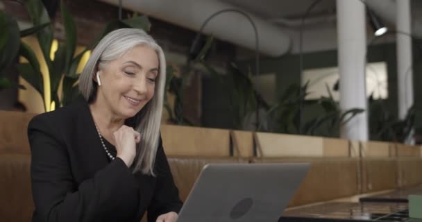 Widok na stronę szczęśliwej nowoczesnej staruszki w formalnym garniturze, mającej wideokonferencję na laptopie. Dojrzałe eleganckie businesswoman za pomocą portative komputer do komunikacji wideo podczas siedzenia w kawiarni. — Wideo stockowe