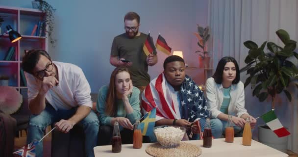 Gruppe multiethnischer Freunde, die gelangweilt auf dem Sofa neben dem Fernseher sitzen. Männliche und weibliche Freunde mit Fahnen verschiedener Länder verfolgen Sportspiele mit Desinteresse . — Stockvideo