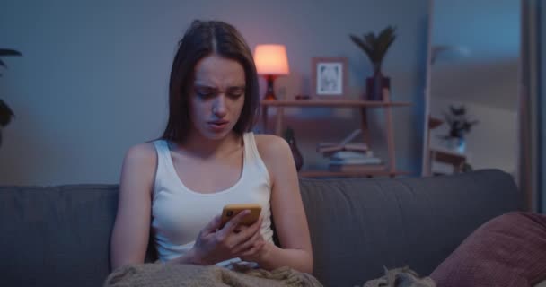 Nešťastná mladá dívka dostává zprávy do postele na svém smartphonu, zatímco sedí na pohovce doma. Ženské tisícileté vypadající ustaraně a smutně při kontrole obsahu mobilního telefonu. Indoors. — Stock video