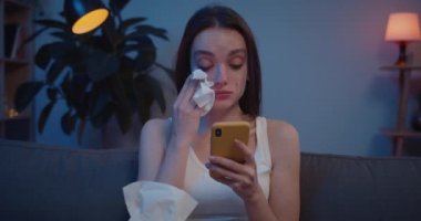 Genç bir kadının telefon ekranına bakarken yüzünü kağıt mendille silişi. Üzgün kız, akıllı telefonunda kötü haberlere ve ağlamaya inanamaz..