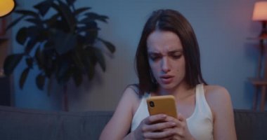 Akıllı telefonuyla kanepede oturan genç bir kadının ekrana bakıp üzüldüğü yakın plan. Güzel kız, ayrılık mesajını okuduktan sonra yüzünü elinle kapatıyor ve ağlıyor. .