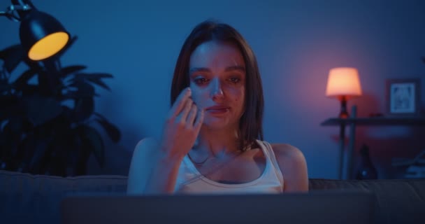 Close up of young upset girl in tears watching drama on her laptop while sitting on sofa. Sad millennial woman crying and wiping tears with paper tissue at home in evening. — Stock Video