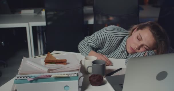 Uykulu genç bayan ofis çalışanı yemek masasında önünde açık bir defterle yatıyor. Boş bir gece ofisinde uzun saatler çalışırken uyuyan yorgun bir kadın. Aşırı çalışma kavramı. — Stok video