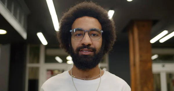 Kameraya bakıp gülümseyen gözlüklü yakışıklı adamın portresi. Modern binanın içinde duran 30 'lu yaşlardaki neşeli sakallı adama yakından bakın. İnsan ve yaşam tarzı kavramı.