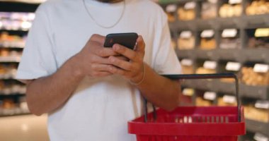 Alışveriş sepetinin yanında duran adamın modern cep telefonunu kullanış görüntüsü. Dükkandan yiyecek alırken akıllı telefondan mesajlaşan ve sohbet eden bir adam..