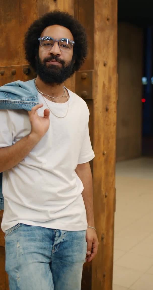 Retrato de un tipo barbudo con ropa casual sosteniendo la chaqueta en su hombro y mirando a la cámara. Hombre maduro en gafas de pie apoyado en la pared. Antecedentes. Vídeo vertical . — Vídeo de stock