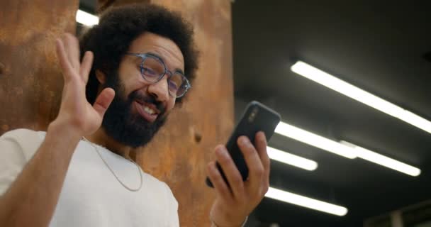 Vista inferiore di ragazzo eccitato negli anni '30 sorridente e agitando durante la conversazione video su smartphone. Uomo in occhiali sembra felice mentre utilizza il suo smartphone per la comunicazione . — Video Stock