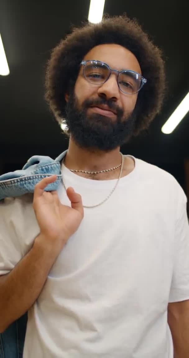 Bottom view of good looking man in white t shirt standing and holding jacket on his shoulder. Portrait of confident guy in glasses looking to camera Vertical video. Loft background. — Stock Video