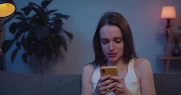 Detailní záběr mladé dívky pláče při pohledu na fotografie se svým přítelem po rozchodu. Smutná mladá žena sedí na pohovce a dívá se na obrazovku smartphonu a pláče po obdržení špatné zprávy . — Stock video