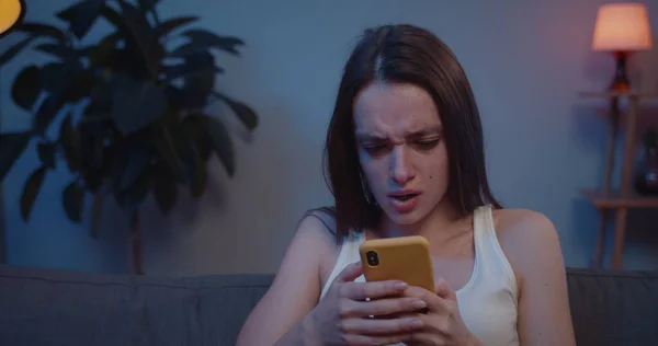 Akıllı telefonuyla kanepede oturan genç bir kadının ekrana bakıp üzüldüğü yakın plan. Güzel kız, ayrılık mesajını okuduktan sonra yüzünü elinle kapatıyor ve ağlıyor. .