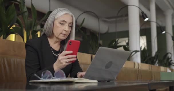 Front view of old woman enterpreneur looking at smartphone screen and typing on her laptop. Successful businesswoman sitting in cafe and working with modern gadgets. Concept of tech and work. — Stock Video