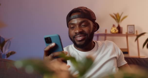 Sorrindo jovem recebendo mensagem engraçada ao usar o smartphone. Bonito homem de boné preto olhando na tela do telefone enquanto se senta no sofá e relaxante. Conceito de lazer . — Vídeo de Stock