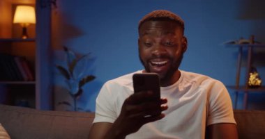 Kanepede oturan ve akıllı telefon kullanarak arkadaşlarıyla iletişim kuran mutlu bir Afrikalı. Heyecanlı görünen genç adamın ön görüntüsü ve iyi bir haber telefonu olduğunda yazmaya başladı..