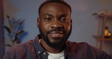 Yakışıklı Afrikalı adamın iyi bir ruh hali içinde kameraya bakışının yakın görüntüsü. Gülümseyen yakışıklı bir adamın portresi. İnsanlar ve duygular kavramı. Rahat ev arkaplanı.