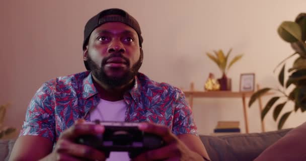 Handsome young man sitting on a couch and using gaming console for playing. Excited man wearing cap looking on TV screen while video gaming at home. Room with neon lights. — Stock Video