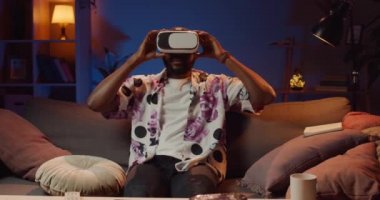 Sanal gerçeklik gözlüğü takan ve etrafı kolaçan eden Afro-Amerikan genç adam. Yakışıklı adam kanepede oturuyor ve evde boş vakit geçirirken kulaklık takıyor. Teknoloji kavramı.