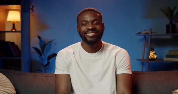 Retrato de hombre feliz en camiseta blanca sonriendo y mirando a la cámara. Vista frontal de chico africano guapo de buen humor sentado en el sofá y mirando feliz. Concepto de ocio . — Vídeo de stock