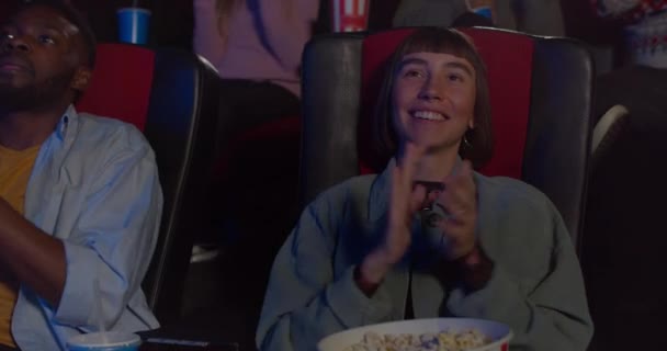 Vista frontal de una joven hipster aplaudiendo y sonriendo después de ver una buena película en el cine. Chica que mira satisfecha con el cine que ve en el cine. Personas de fondo . — Vídeo de stock