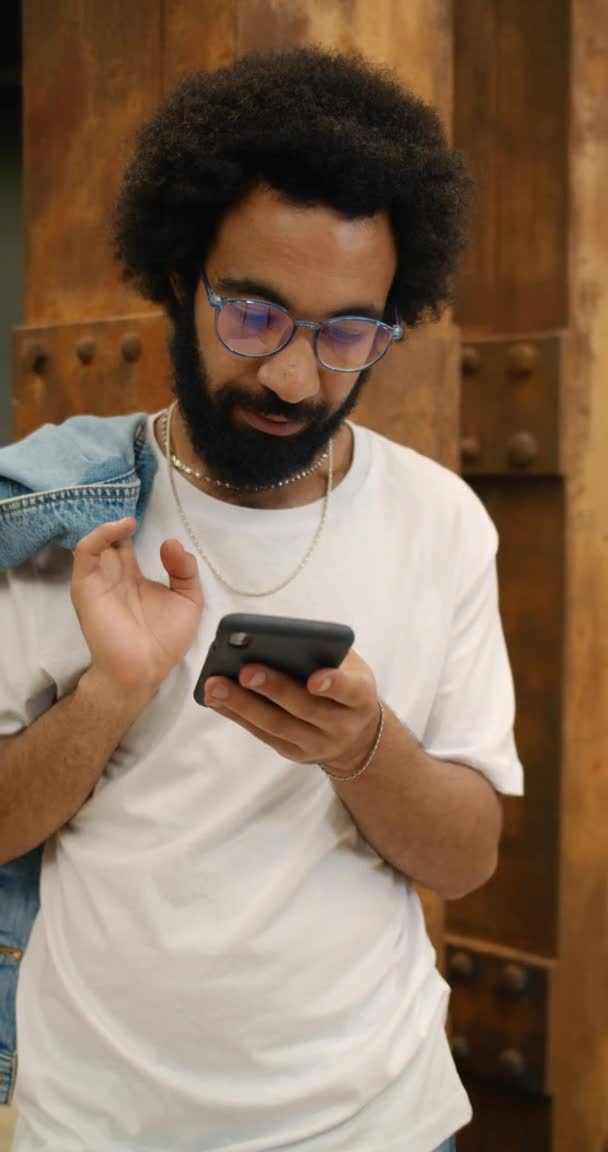 Um tipo sério nos anos 30 a usar o smartphone enquanto estava de pé encostado à parede. Homem de óculos e t-shirt branca necessidades de rolagem alimentar e segurando jaqueta no ombro. Vídeo vertical . — Vídeo de Stock