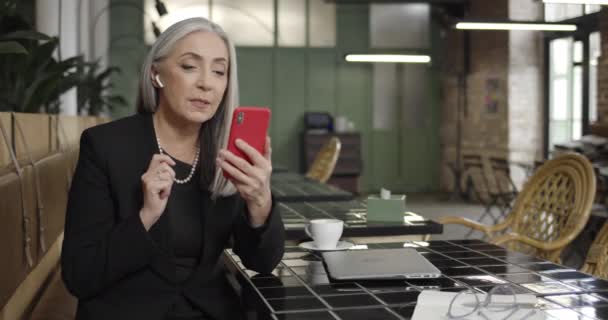 Widok z przodu dojrzałej pani przedsiębiorca w słuchawkach posiadające videocall na smartfonie z partnerem biznesowym. kobieta w średnim wieku komunikujących, za pomocą aplikacji wideo podczas siedzenia w kawiarni . — Wideo stockowe