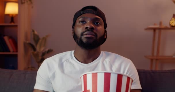 Primer plano de chico guapo agitando la cabeza y empezando a usar el teléfono inteligente mientras ve la película. Hombre barbudo con gorra sosteniendo palomitas de maíz y sonriendo mientras está sentado en el sofá delante de la TV . — Vídeos de Stock