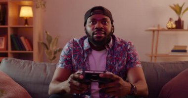 Şapkalı, video oyunu oynayan, evdeki kanepede otururken konsol kullanan heyecanlı bir Afrikalı. Boş zaman geçirirken elinde kablosuz kontrolör tutan tişörtlü mutlu adamın ön görüntüsü.