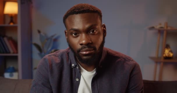 Portrait de jeune homme barbu en vêtements décontractés regardant à la caméra. Gros plan d'un beau mec avec un visage sérieux assis sur le canapé et levant les yeux vers la caméra. Cosy fond de maison . — Video