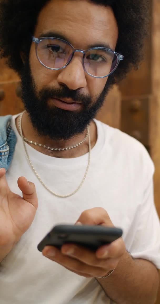 Primer plano de chico en gafas usando smartphone y sonriendo a la cámara. Feliz hombre barbudo sosteniendo su teléfono y levantando los ojos a la cámara mientras estaba de pie apoyado en la pared. Vídeo vertical . — Vídeos de Stock