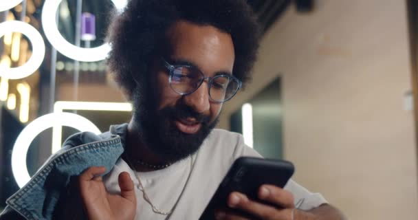 Vue rapprochée de l'homme mûr utilisant son smartphone et souriant tout en se tenant dans un café. gai barbu guy dans lunettes regardant joyeusement écran de téléphone et tenant veste sur son épaule . — Video