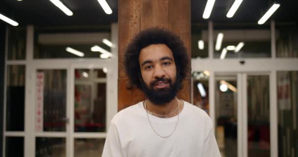 Portret van een vrolijke man in wit t-shirt kijkend naar de camera en glimlachend. Baard man in de 30 staat in een modern gebouw. Concept van mensen en levensstijl. Loft achtergrond. — Stockvideo