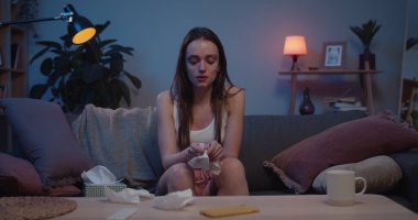 Kanepede oturan ve elinde kağıt mendil tutan mutsuz bir kızın ön görüntüsü. Depresyondaki genç bir kadın ağlıyor ve kameraya bakıyor. Ev geçmişi. Duygular ve duygular kavramı.